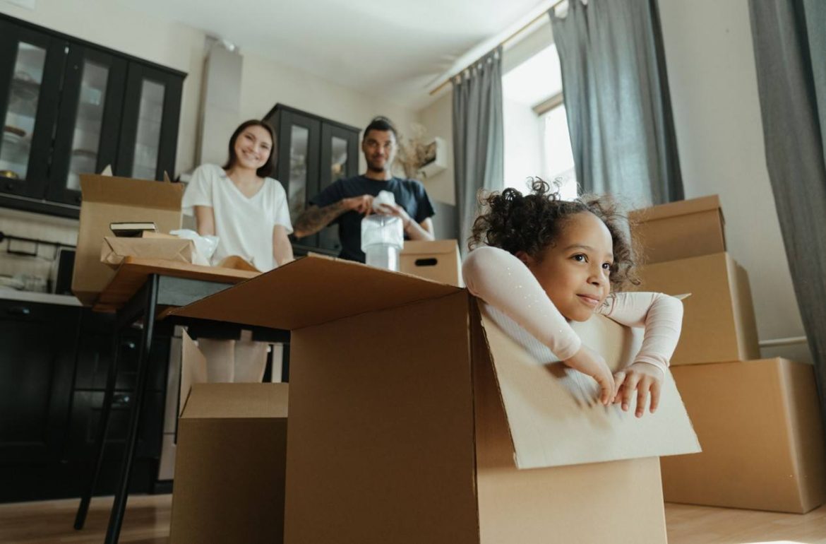 Une petite fille sourit à l'intérieur d'un carton, illustrant les astuces face au déménagement enfants stress alors que ses parents s'activent en arrière-plan.