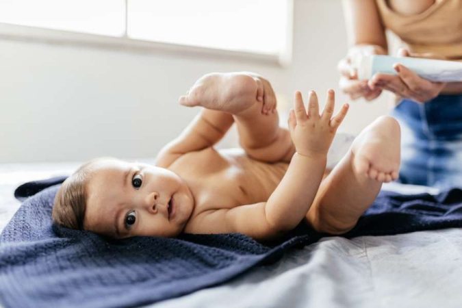 Un bébé allongé sur le dos regarde l'objectif pendant que sa mère tient une crème contre l'érythème fessier en arrière-plan.